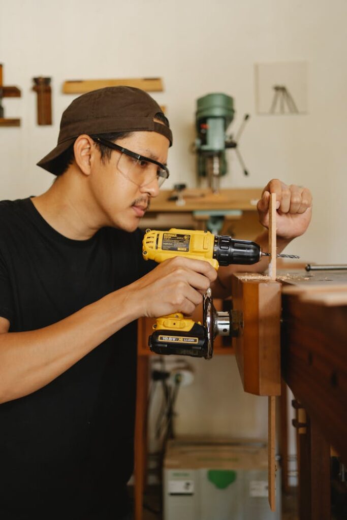 Side view of ethnic male in cap drilling timber plank with screwdriver in workshop with equipment